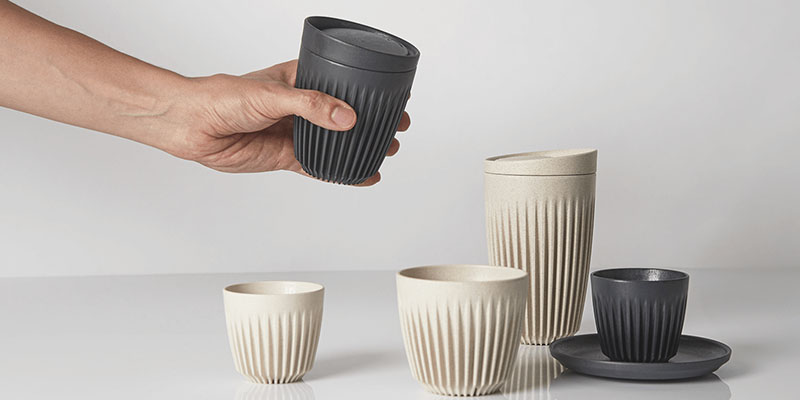 A male hand holds a black Huskee coffee cup in the air. A mix of neutral and black  coffee cups rest below on a bench.