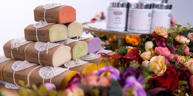 An image of a neatly stacked pile of handmade goat milk soap, each bar wrapped in brown paper and tied with lace and string. In the foreground are brightly coloured flowers. The background is a blurred image of white and silver jars of beauty products.