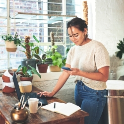 A person uses a phone and laptop at a workbench in a studio filled with ceramics and plants.