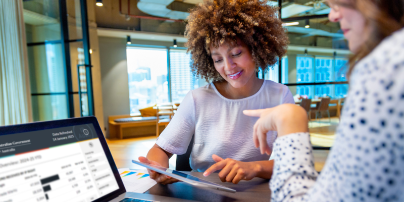 Two women discuss a project at a table, one has a laptop in front of her with the other referencing an iPad