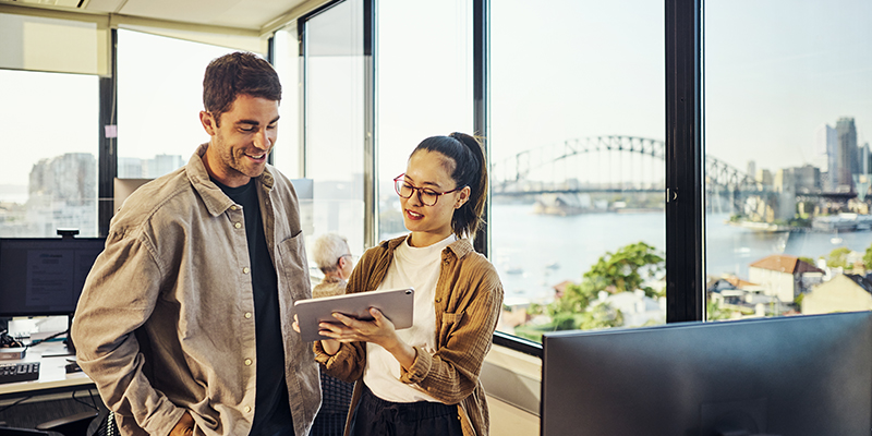 A male and female look at an ipad in an office building with the Sydney Harbour bride in the distance.