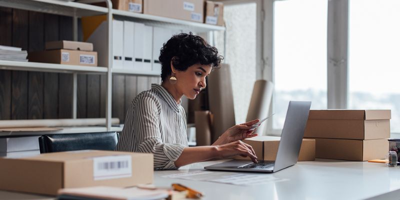 A well dressed woman of colour checks her laptop in an office filled with boxes and files on shelves.