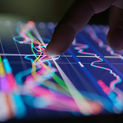 A person uses their finger to interact with a tablet displaying colourful financial data charts and trend lines on a dark screen.