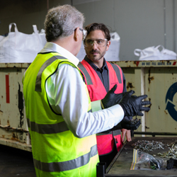 Two people wearing high-visibility safety vests and protective gloves discussing materials at a recycling facility.